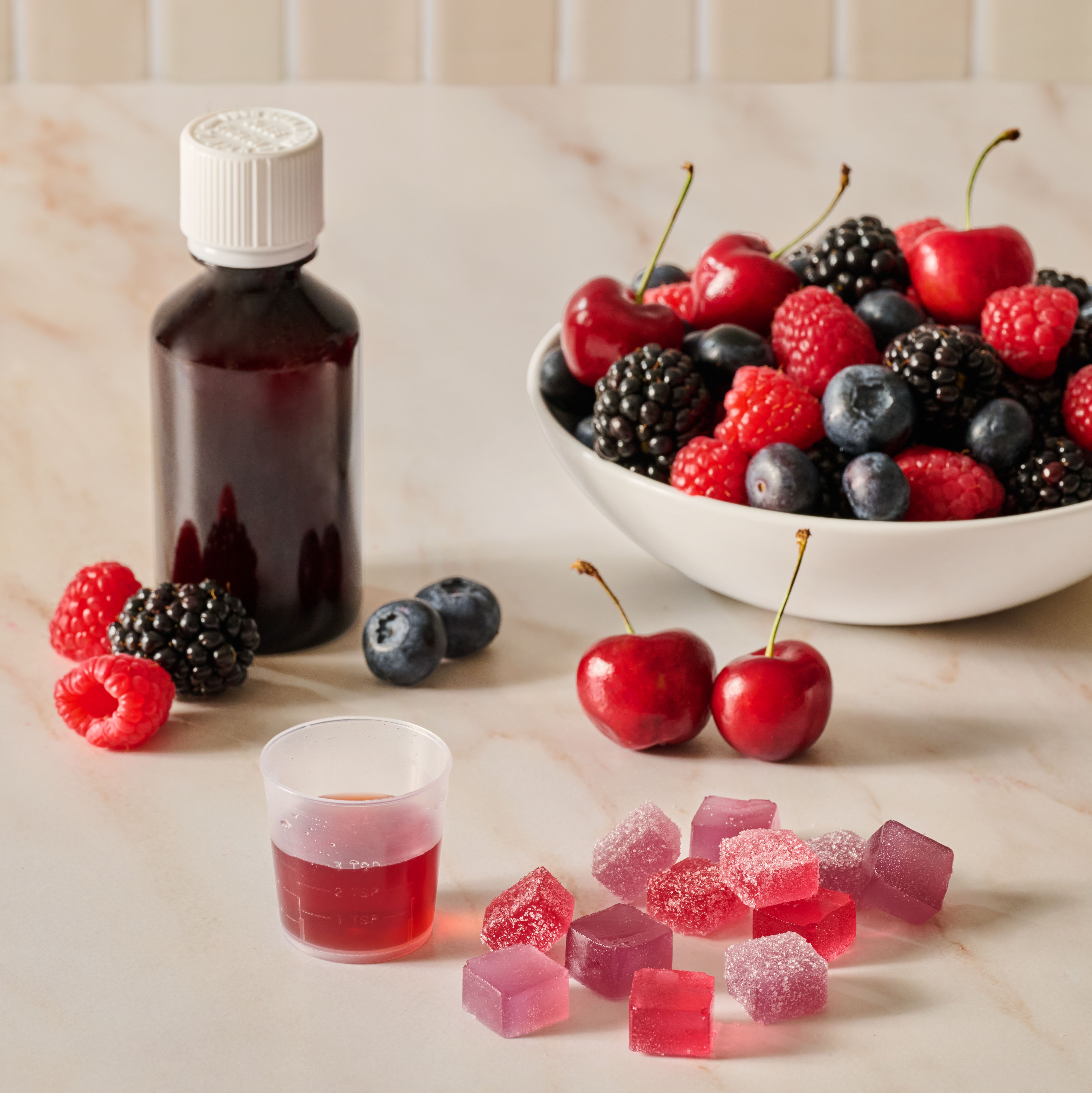Image of syrup medicine in a small cup and gummies in front of a bowl of cherries and berries.