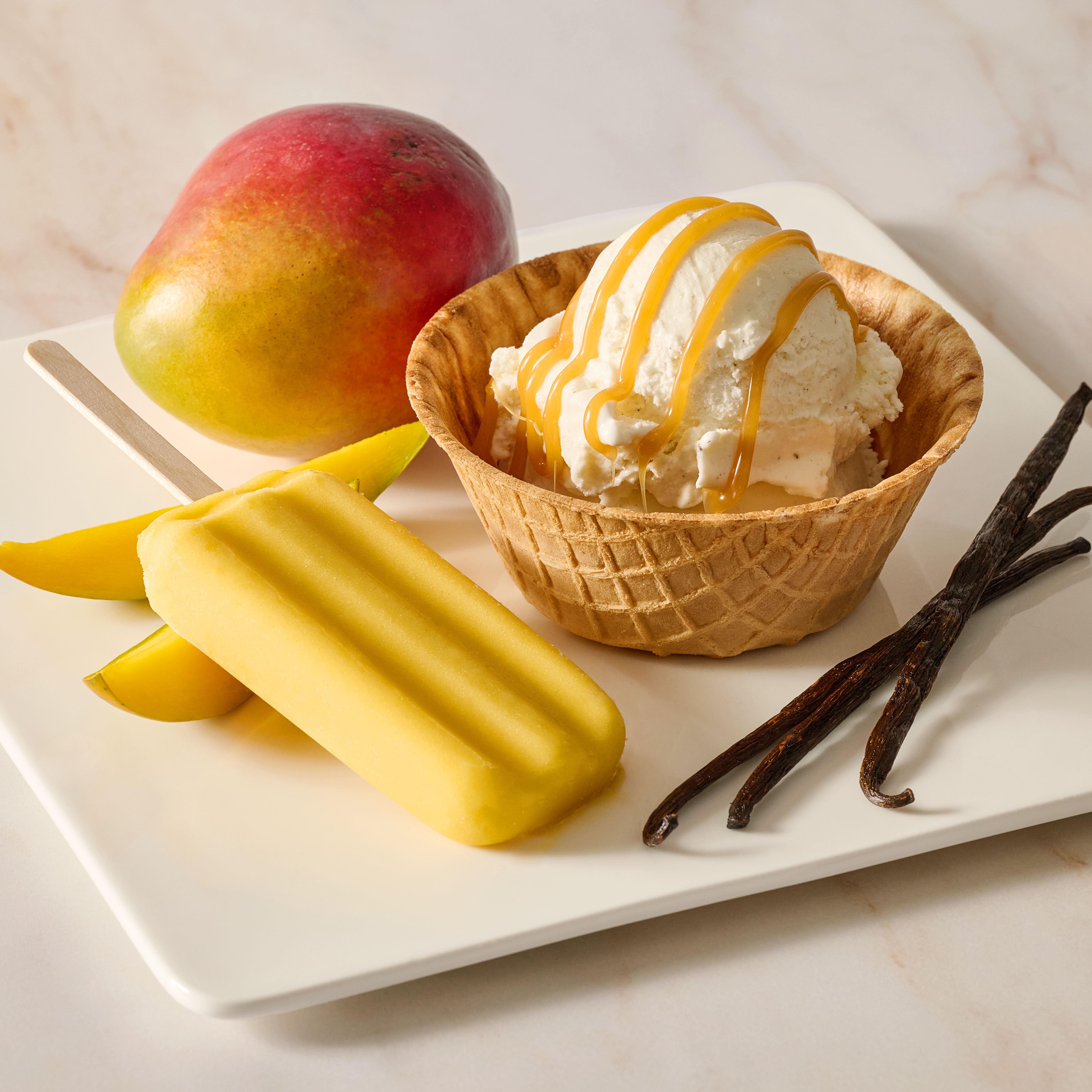 Image of a popsicle and ice cream in a waffle bowl with a mango and vanilla beside it.