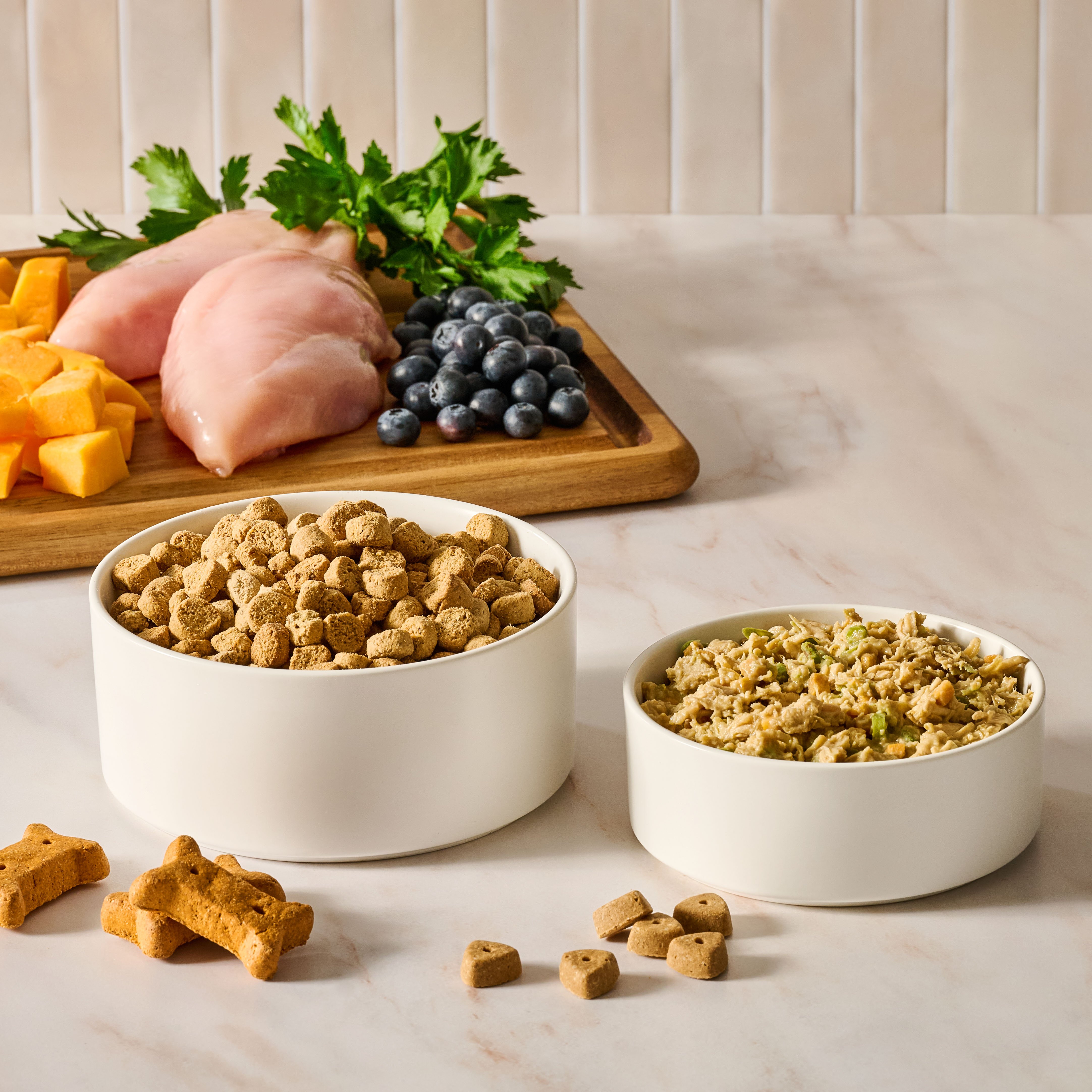 Bowls of dry and wet pet food with ingredients such as poultry, fruits, and vegetables behind them.