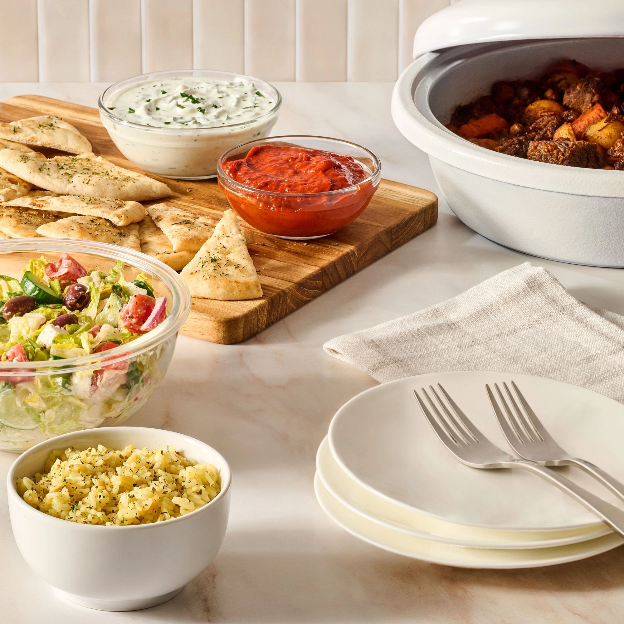 Tablesetting with roasted meat, pita and dips, salad, and seasoned rice with plates and silverware next to it.